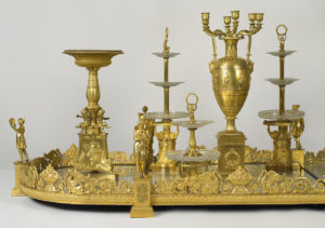 A gilt-bronze table centerpiece with mirrored glass and hand engraving comprised of classical figures, 3-tiered serving dishes, and Classical ornamental figures and motifs, dated to 1805 and on view in Tablescapes: Designs for Dining.
