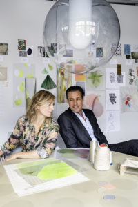 Design by Hand. Image of a woman and man seated at table in front of pictures, and materials samples attached to a wall in the background.
