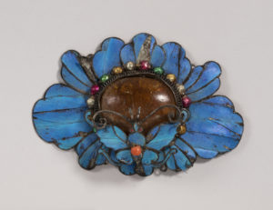 Image features an ornament in the form of a butterfly alighting on a blossom, depicted in bright blue kingfisher feathers, brown resin, and red and green foil-covered beads mounted on a sheet silver backing. Please scroll down to read the blog post about this object.