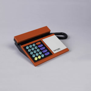 Image features rectilinear desktop telephone in red and black, with handset at top, black keypad with green, yellow and blue number and function keys on left, rectangular gray panel with logo on right. Please scroll down to read the blog post about this object.