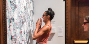 A woman with her hair in a top knot and dressed a pink tank top wearing 3D glasses smiles broadly as she views an installation of floral wallpaper that appears 3 dimensional when wearing the glasses Scroll down for information on the museum's current exhibitions.