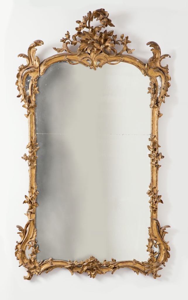 Image features Mirror in large, carved and gilded vertical frame with double arch at top with pyramid of asymmetrical and tall curved leaf forms at each corner which flow into serpentine curves and then join straight sides; flowers entwined around straight sides; bottom rail duplicates curve at top rail and has asymmetrical group of foliage carved at center. Please scroll down to read the blog post about this object.