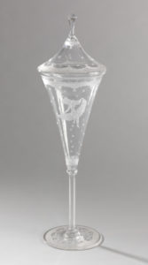Image features vessel with longated conical shaped bowl; bowl has engraved decoration of female figure sitting on ruffled banner, scalloped ornamental bands at top and bottom and stars spaced throughout. Tall, thin six-sided stem with small notches on corners. Flat circular foot with ornamental bands that match bowl. Lid tapers to tall six-sided notched finial with knop. Please scroll down to read the blog post about this object.