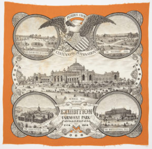 Commemorative handkerchief made for the Centennial Exhibition of 1876. In the center is the Memorial Hall Art Gallery surrounded by roundels containing the Agricultural Hall, Horticultural Hall, Machinery Hall, and the Main Exhibition Building. Between the two top roundels is an eagle with a shield flag and a banner clasped in its beak that reads “E Pluribus Unum.” Border is orange while the exhibition buildings are black and white.