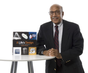 A photograph of industrial designer Charles Harrison, winner of the 2008 National Design Award for Lifetime Achievement. Harrison wears a suit and tie and is seated next to white round table on which there is a copy of a biography of the designer.