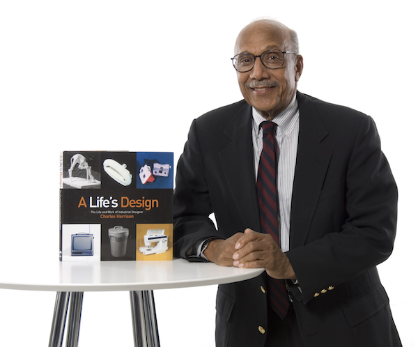 A photograph of industrial designer Charles Harrison, winner of the 2008 National Design Award for Lifetime Achievement. Harrison wears a suit and tie and is seated next to white round table on which there is a copy of a biography of the designer.