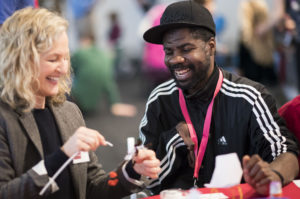 Image of a young black man with Cerebral Palsy seated next to an older white woman. They work together on a paper prototype, both are smiling.