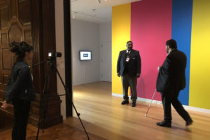 Image of accessibility consultant Sina Bahram in front of a colorful wall in the Cooper Hewitt Galleries. Sina walks towards a museum security guard, in front of a woman who records their interaction with a camera.
