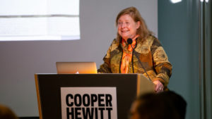 Sarah Coffin, a blond woman in her 60s stands at a podium that has the Cooper Hewitt sign. She wears a colorful scarf.