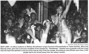 A black and white photograph of Taana Gardner performing, surrounded by a densely packed crowd. She sings into a microphone with one hand raised, wearing a light-colored, strapless dress. The crowd surrounding her is comprised mostly of people of color.