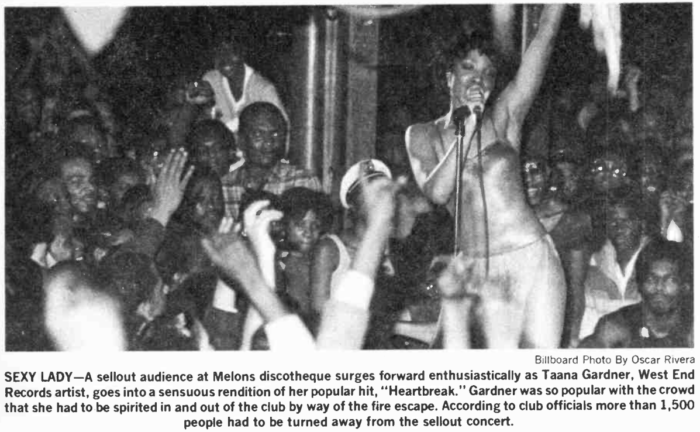 A black and white photograph of Taana Gardner performing, surrounded by a densely packed crowd comprised mostly of people of color. She sings into a microphone with one hand raised, wearing a strapless dress.
