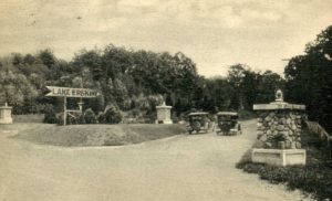 Horizontal black-and-white historical photograph of an intersection of two streets. The perspective is from the street in the foreground (moving across the image left to right) looking down the intersecting street. Two cars are stationed down the intersecting streets, and along the size of the road are, on the left, a wooden, arrow-shaped sign that reads Lake Erskine and on the left a stone pier with what appears to be a small lantern on top of it. The background of the photo is lined with trees and the sky above is partly cloudy.