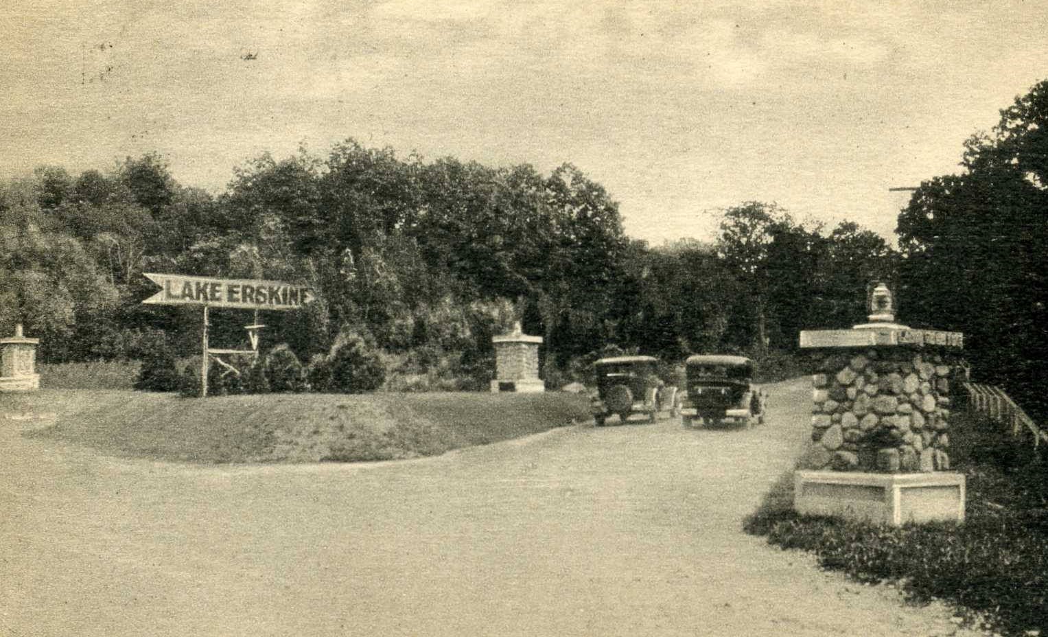 Horizontal black-and-white historical photograph of an intersection of two streets. The perspective is from the street in the foreground (moving across the image left to right) looking down the intersecting street. Two cars are stationed down the intersecting streets, and along the size of the road are, on the left, a wooden, arrow-shaped sign that reads Lake Erskine and on the left a stone pier with what appears to be a small lantern on top of it. The background of the photo is lined with trees and the sky above is partly cloudy.