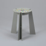 A grey thermoplastic 3-legged stool with a circular seat featuring a multicolor pattern of small squares.