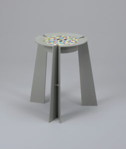 A grey thermoplastic 3-legged stool with a circular seat featuring a multicolor pattern of small squares.