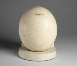Image features a large egg carved with the allegorical image of "America" as a female figure wearing a robe and feather headdress, holding arrows, and seated before a palm tree and crocodile within a landscape. Please scroll down to read the blog post about this object.