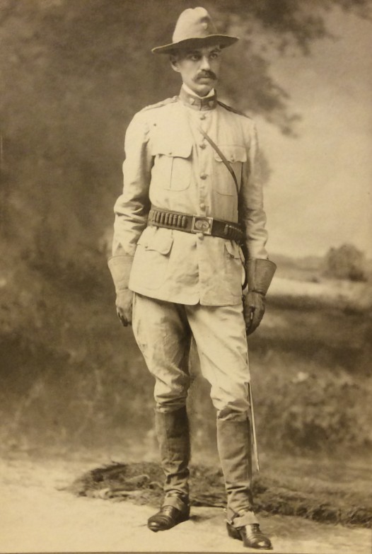 A black-and-white portrait of a tall man in millary uniform. He stands are center against a background of trees and sky, looking to the right of the viewer with his left foot positioned slightly out in front of himself. He wears a wide-brimmed hat, an tan jacket with a high collar and many pockets, gloves, tan pants, and boots.