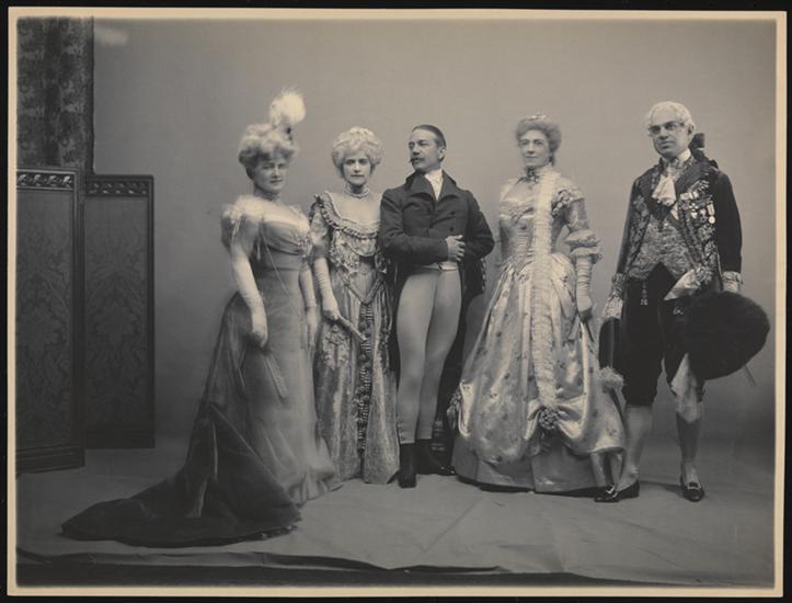 A black-and-white historical photograph of five people. They are all standing in pose against a blank background, and they are dressed in elaborate costume. The costumes are a mix of historical styles.