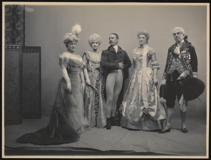 A black-and-white historical photograph of five people. They are all standing in pose against a blank background, and they are dressed in elaborate costume. The costumes are a mix of historical styles.