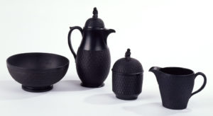 Photograph of four stoneware vessels. From left to right: a bowl that is circular, on raised foot, sides have basket-weave texture, inside smooth. Next a chocolate pot that is tall, slightly swelling body, long loop handle, short spout; high domed lid with finial in the form of a draped female figure. Sides of body (lower portion) and lid with basket-weave texture; wide band top section of body plain. Third, a sugar bowl that is cylindrical, high domed lid with finial in the form of a draped female figure; basket-weave texture sides and lid. Finally, a creamer that is a slightly swelling form, short spout, long loop handle; sides with basket-weave texture.
