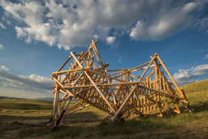 Three-dimensional wooden sculpture set against background of sky and grassy land.