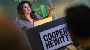 Image of Sarah Williams, Director, Civic Data Design Lab at MIT behind a podium with a Cooper Hewitt sign. She has curly brown hair, a round face, and her hands are raise slightly in front of her as she gestures during a recent talk at Cooper Hewitt