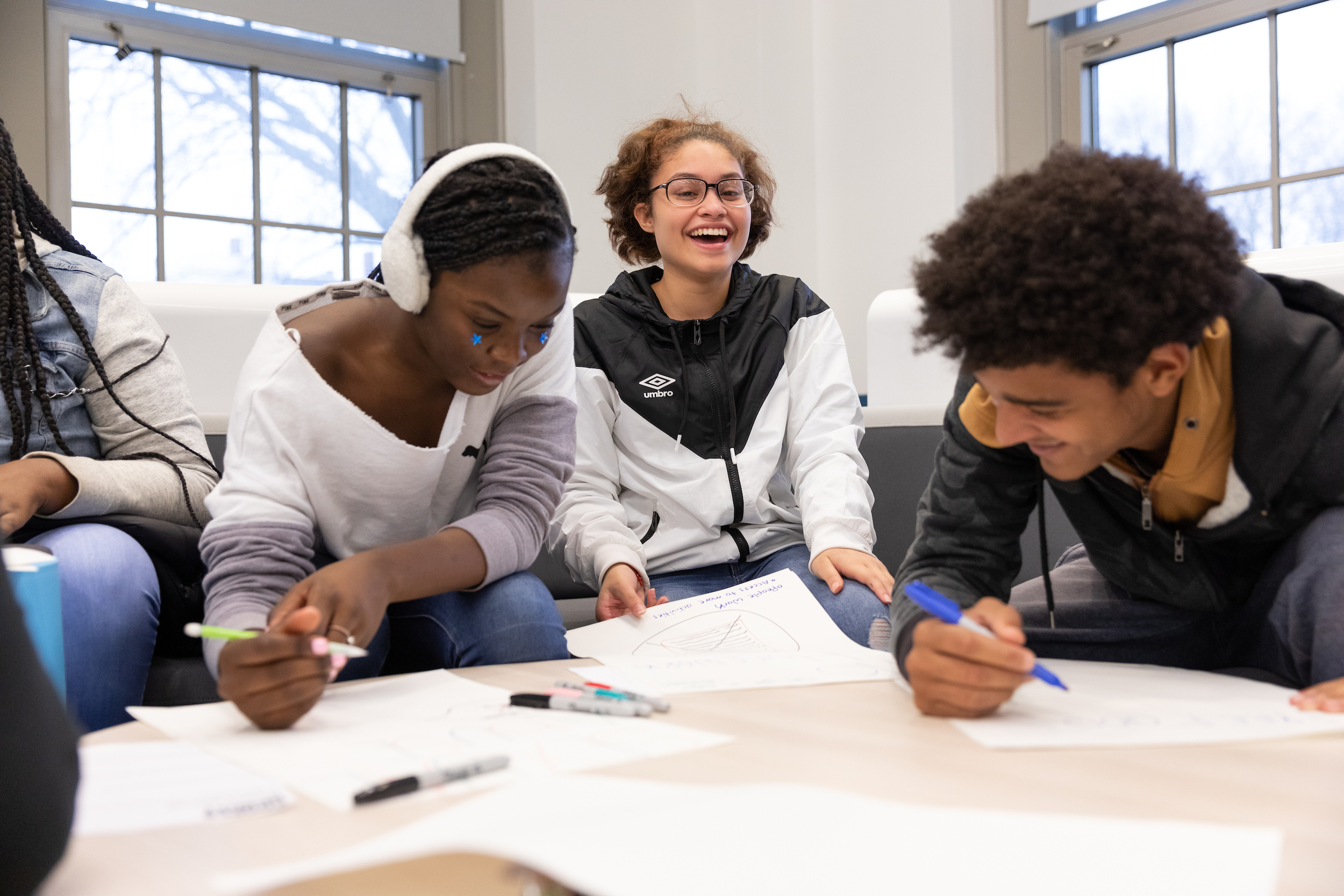 Three young people sit together and smile, two of them draw on sheets of white paper