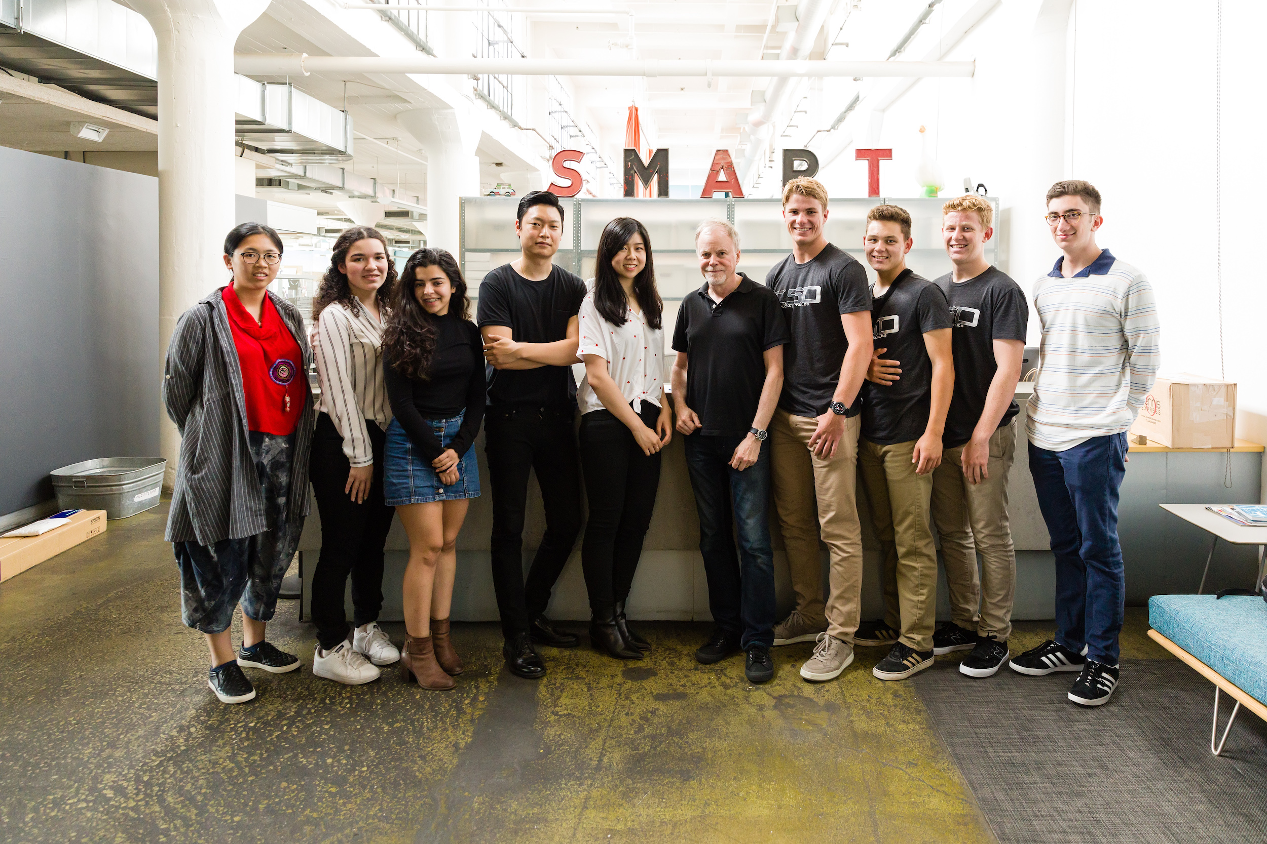 Ten people stand together and smile for the camera in a large, bright room with high ceilings