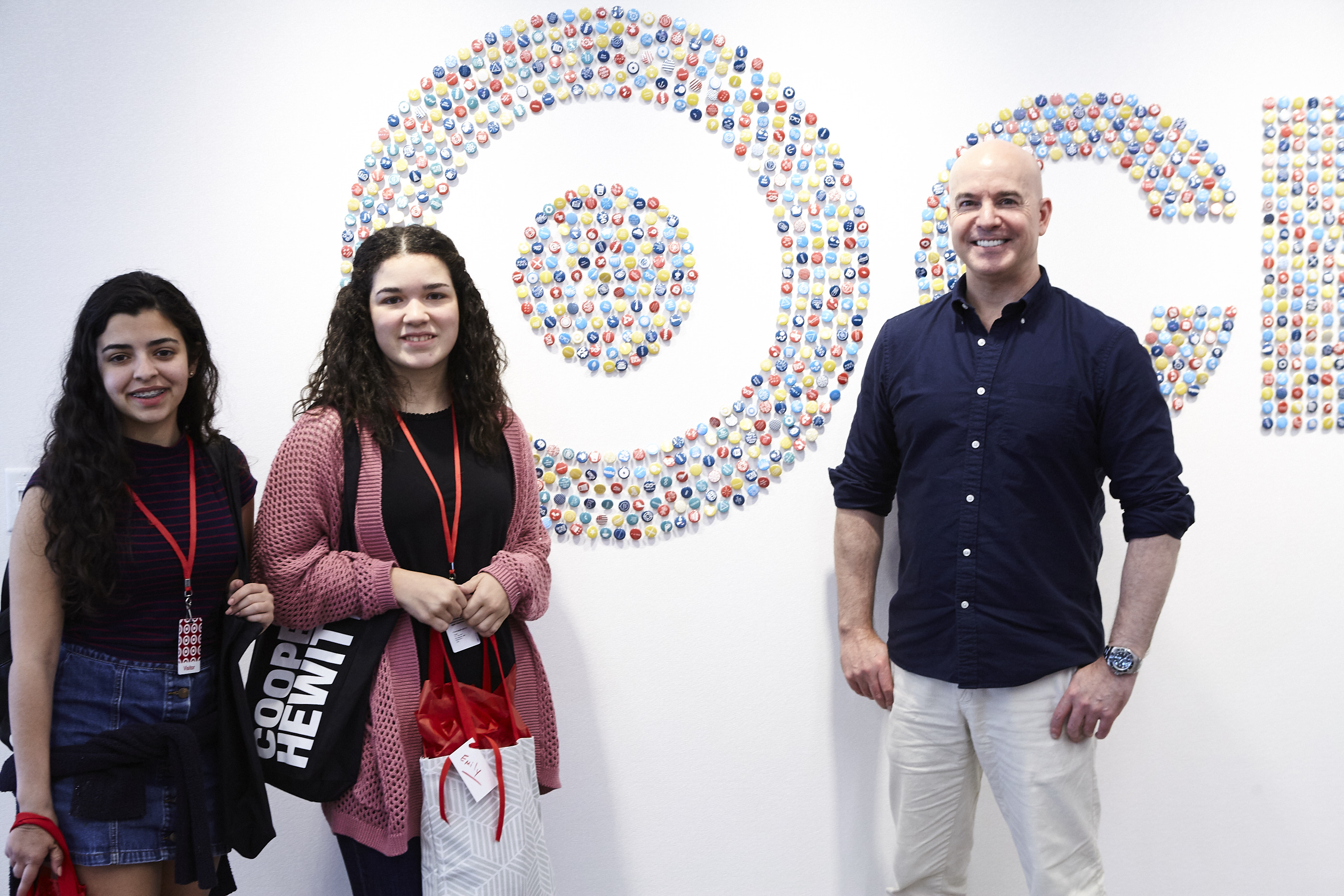 Two young women stand with a man in front of a white wall with a target on it made up of small, colorful circles