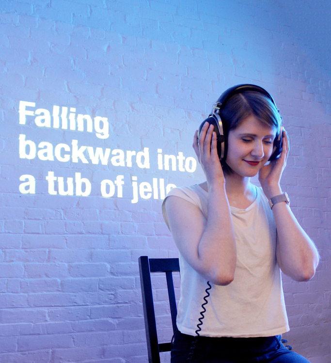 A woman sits with headphones on and her eyes closed, the words Falling backward into a tub of jello are projected on the wall behind her