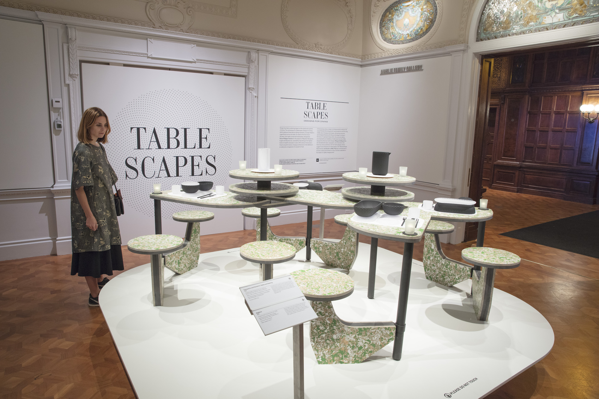 A woman examines a table design which features many connected seats and surfaces. The piece is within the Tablescapes: Designs for Dining exhibit