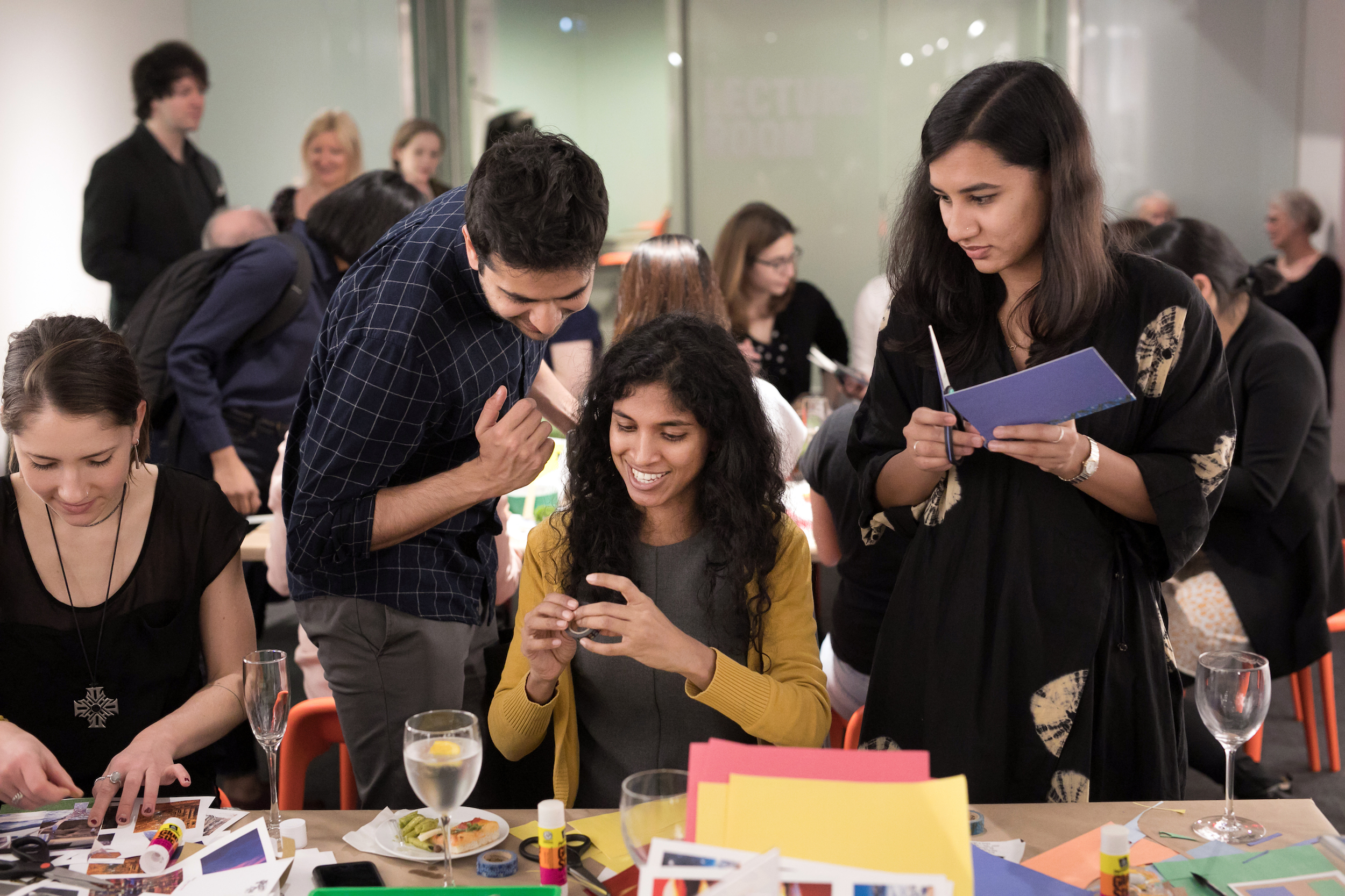 Two standing people interact with a seated woman, who is working with crafting materials on the table in front of her