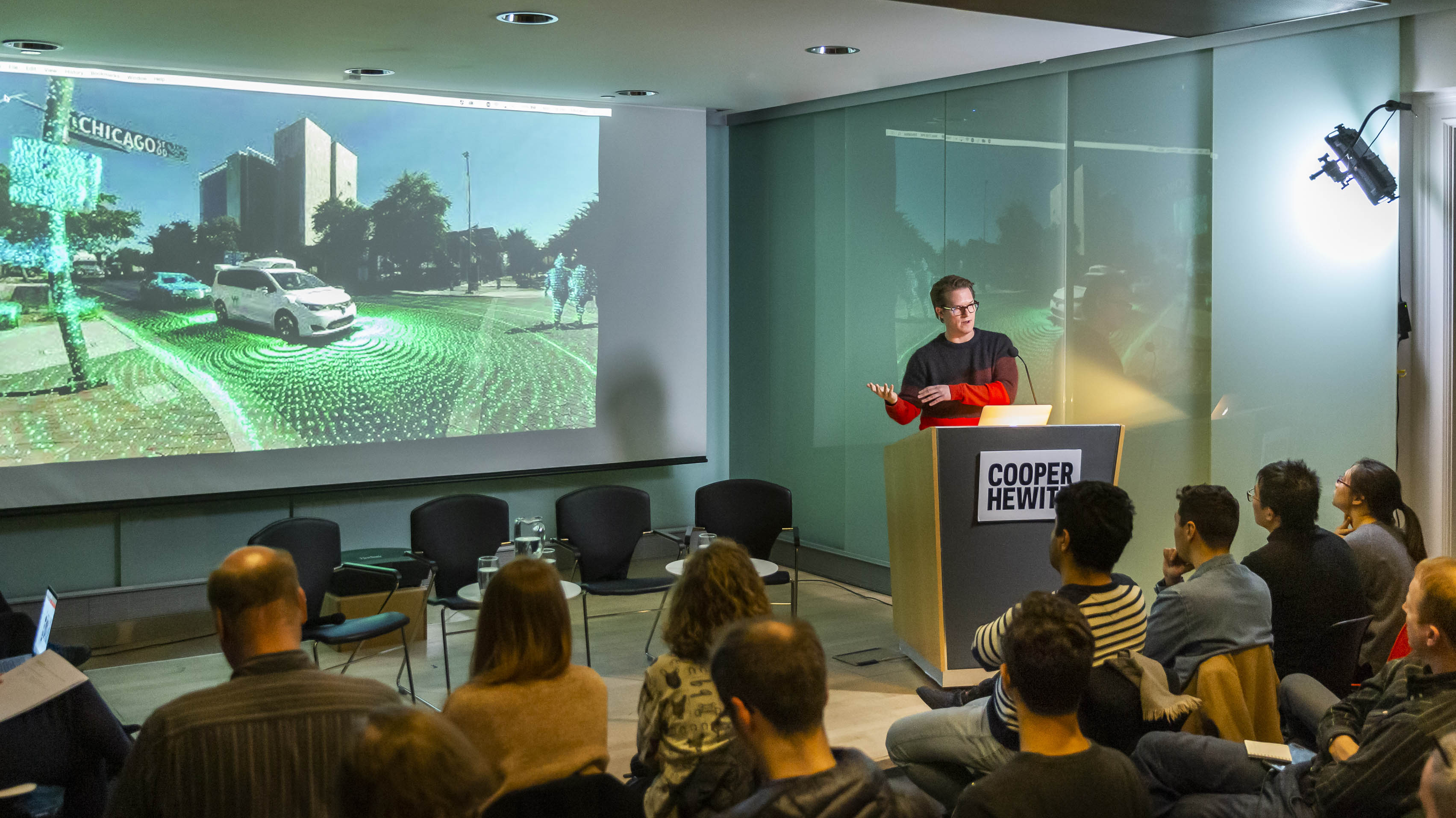 Image of Ryan Powell, head of research and UX design, Waymo behind a podium with a Cooper Hewitt sign. He has brown hair and glasses, and wears a red and blue striped sweater. His hands are raise slightly, gesturing to an image on a screen behind him. The crowd looks on.
