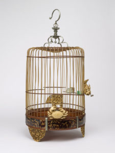 An empty birdcage. The birdcage has bars made of bamboo and a base made of beautiful tortoiseshell. Decorating the bars is an ivory crab.