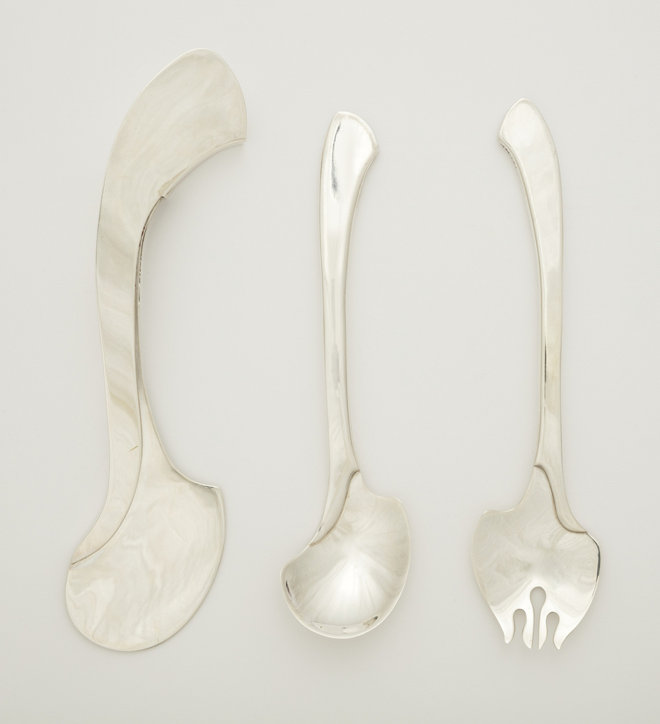 Image features silver portable flatware set of fork, spoon and knife with serpentine outline; flat curving handles leading to curved broad heads and knife blade; Knife handle with two slots to accept, the fork and spoon. Please scroll down to read the blog post about this object.