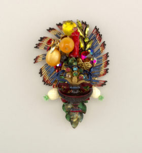Image features brooch of inverted ovoid form composed of various media and costume jewelry fragments: colorful cast metal fringe-like surround, small red figure of Venus, plastic globes suggesting oranges, colored beads and glass pastes, central metal fleur-de-lys, plastic white pineapples, and glass leaves. Please scroll down to read the blog post about this object.