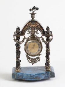 A beautiful ornate historic clock. Mounted on elegant blue marble stone is a miniature trellis. Hanging from the center of the trellis, like a pendant on a necklace, is a clock with roman numerals. The clock is draped with metal and jewels in the form of flowing ribbons and flowering vines embellished with diamonds and pink gemstones. Two small metal owls are perched on either side of the trellis, and on top of the trellis is a small metal vase with metal flowers growing in it. The center of the flowers are pink gemstones.
