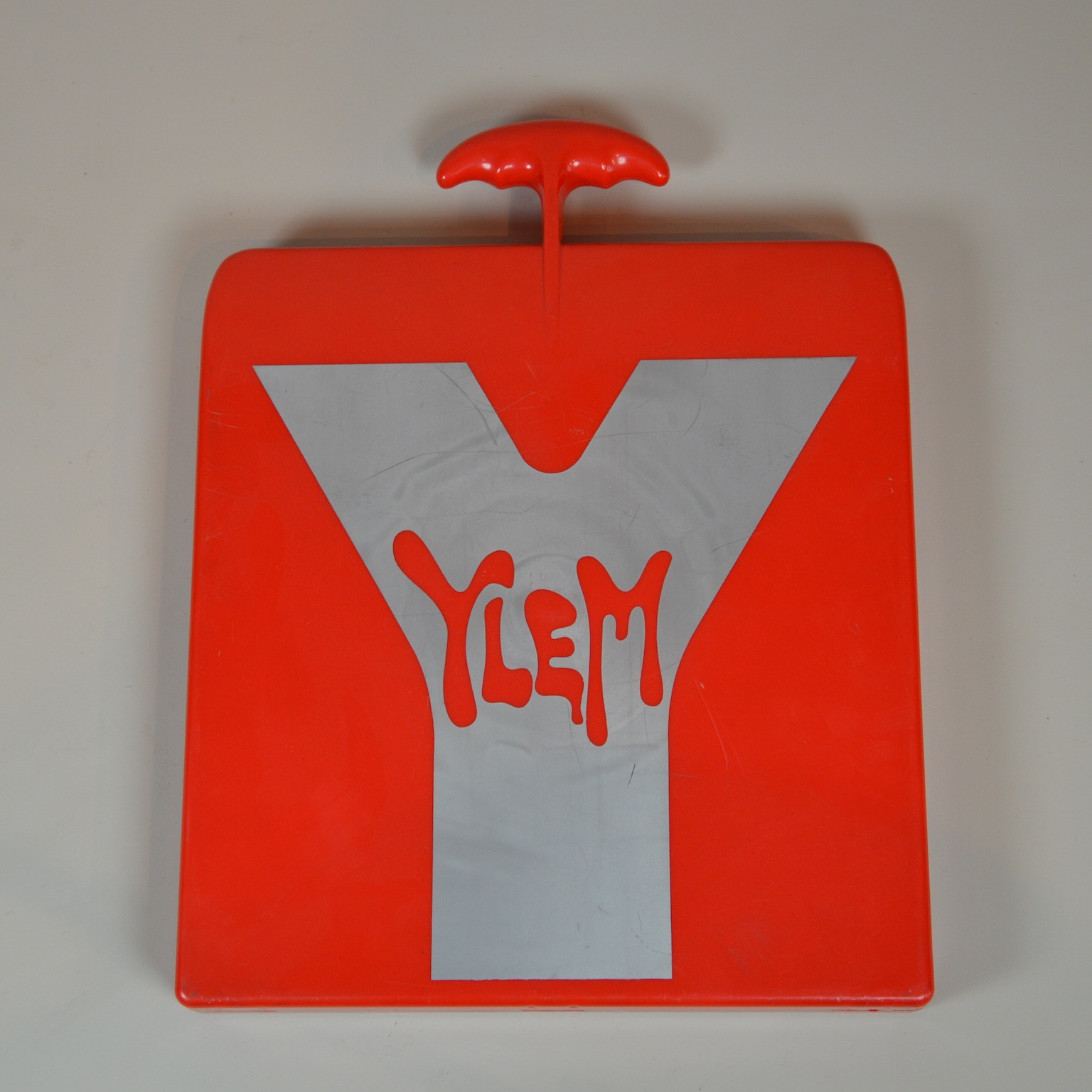 Image features a roughly square book in the form of a bright red plastic portfolio with a T-shaped handle at the top. The cover is embellished with a large silver-toned letter Y with the word YLEM superimposed in red. Please scroll down to read the blog post about this object.