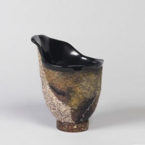 A vessel with an asymmetric form. The body of the vessel is made from a fuzzy material in earthy tones. The inside of the vessel is shiny black.