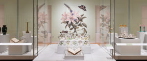 In the galleries of Cooper Hewitt is displayed a large, old fashioned book with its pages open. Behind the book is a case with a set of 12 porcelain plates with botanical models painted on them. Behind the case, printed in jumbo scale on the wall, is a botanical illustration of a flowering plant with pink flowers. Two butterflies with black and yellow wings flutter around the flower.