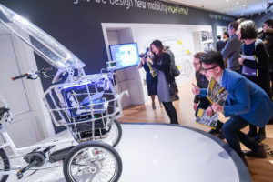 A group of young people in the galleries of Cooper Hewitt interact with the PEV, a robot that looks like a grocery cart combined with a tricycle with glowing blue eyes. The visitors are giving the robot "Thumbs Up" and smiling and laughing.
