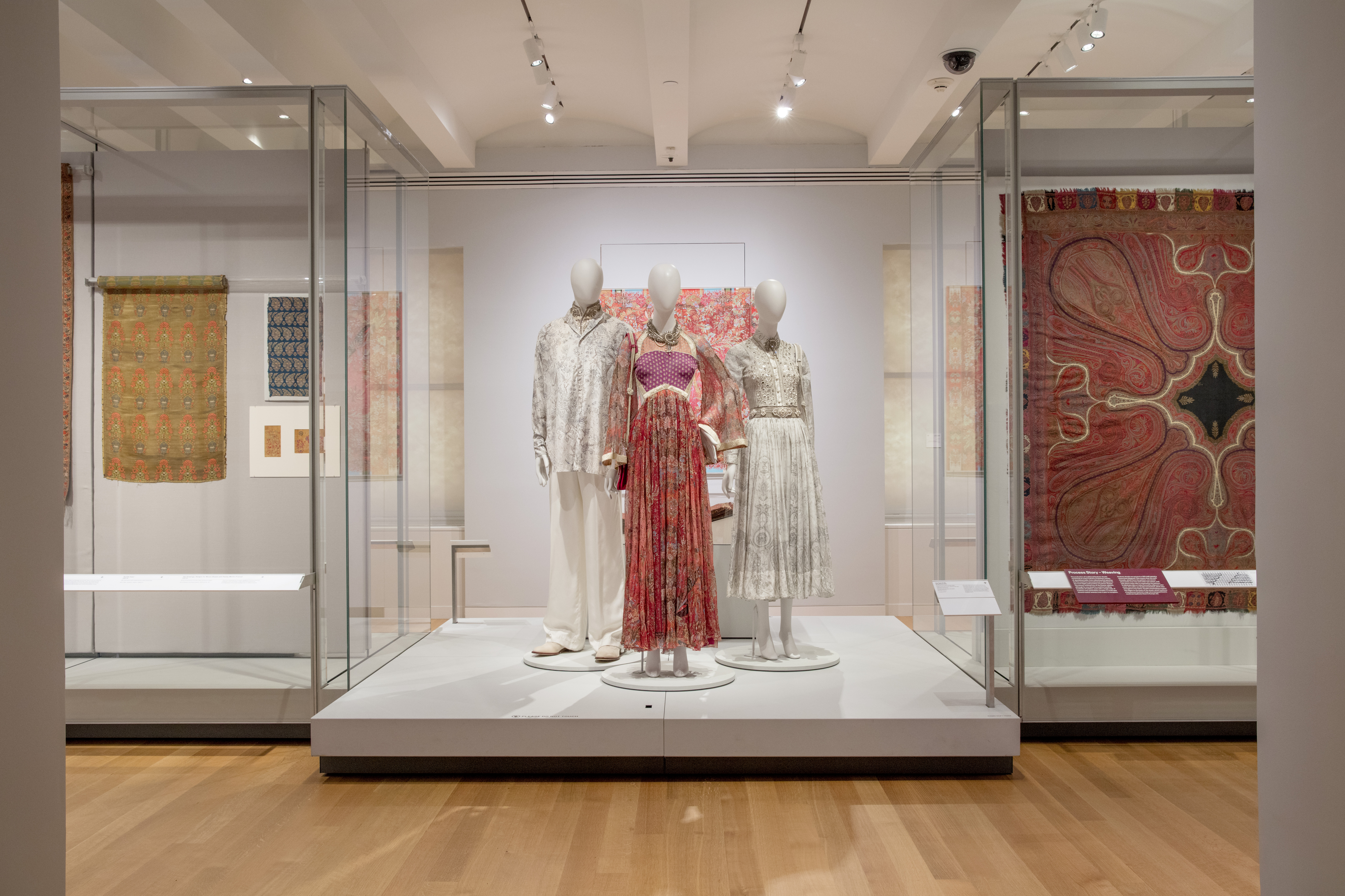 In the Cooper Hewitt galleries, three white mannequins are positioned on a platform. The mannequin the center wears a red gown. The mannequin on the left wears a white silk blouse and white silk pajama pants. The mannequin on the right wears a white gown. To the sides of the mannequins are cases, through which can be seen an enormous red flower fabric hanging.