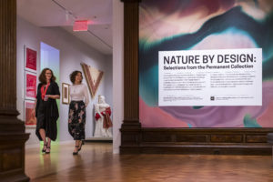 In the galleries of Cooper Hewitt, two well-dressed women stroll past a wall with paisley bandanas hanging on them and toward a wall that says "Nature by Design" decorated with waves of blue, pink and peach tones