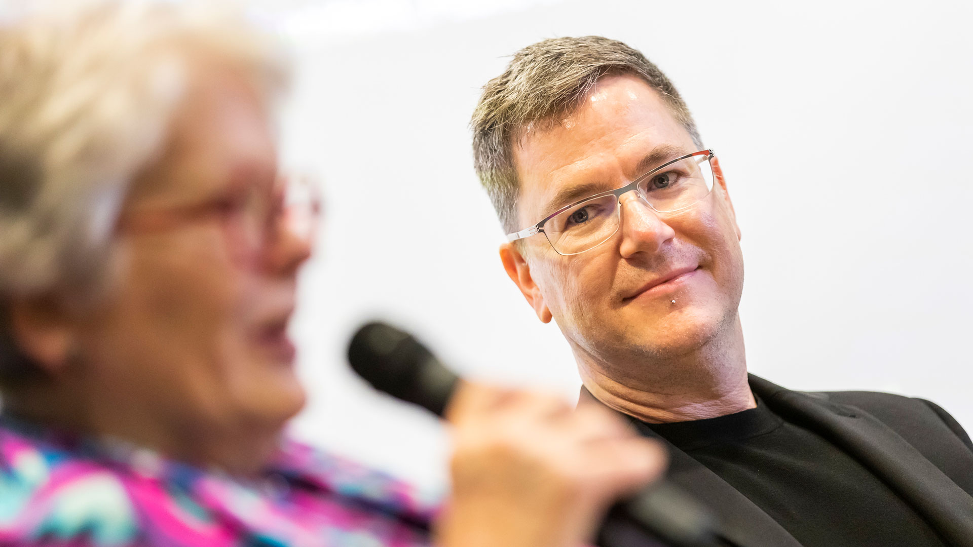 image of two people's heads, one in focus on the right, the other on the left is out of the focus. the one on the right is looking at the one on the left, who holds a microphone to her mouth and appears to be talking. the woman on the left has curly hair and red glasses, the man on the right has short straight grey/brown hair and dark glasses