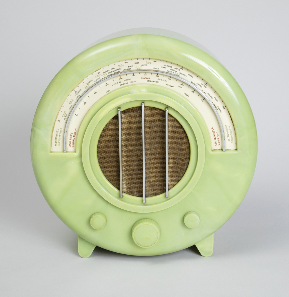 Image features Circular green radio with semi-circular station dial at top front above circular brown textile-covered speaker with three vertical metal rods as grille; three circular control knobs below speaker. Please scroll down to read the blog post about this object.