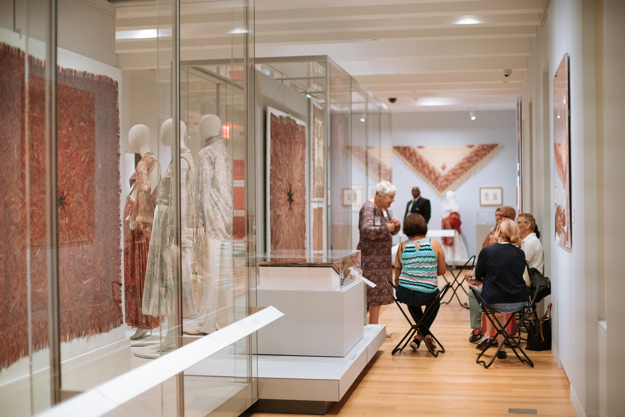 A group of several people seated on stools in a well-lit gallery space a series of red textiles are installed in glass cases beside them