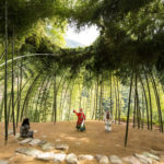 A theater with an earthen ground, rock benches, and a ceiling made of enormous stalks of bent bamboo leaning toward the center of the stage. A man in vibrant red traditional garb gestures as one onlooker sits on a stone.