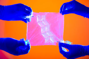 Against a vibrant orange background, four hands in blue gloves tug at the corners of a pink square of plastic that has eight opaque cells in the center, arranged into a zig zag pattern