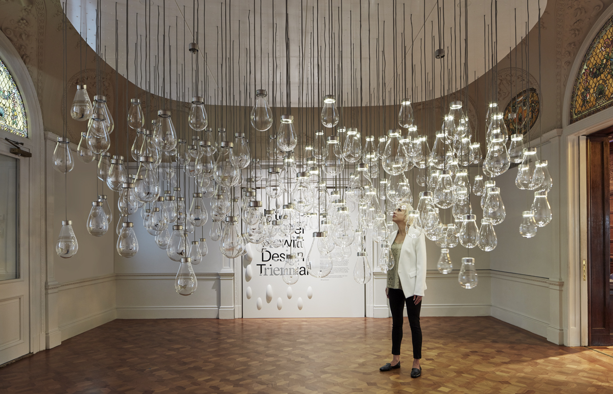 A woman with a blonde bob wearing a white jacked is surrounded by oversized incandescent lightbulbs that hang from the ceiling by strings. She is in the galleries of Cooper Hewitt.