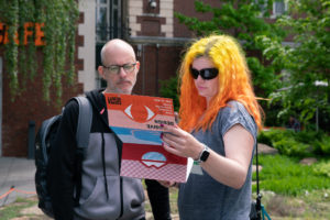 Two people read an oversized colorful brochure with the title "INCLUSIVE DESIGN" while standing in the Cooper Hewitt garden